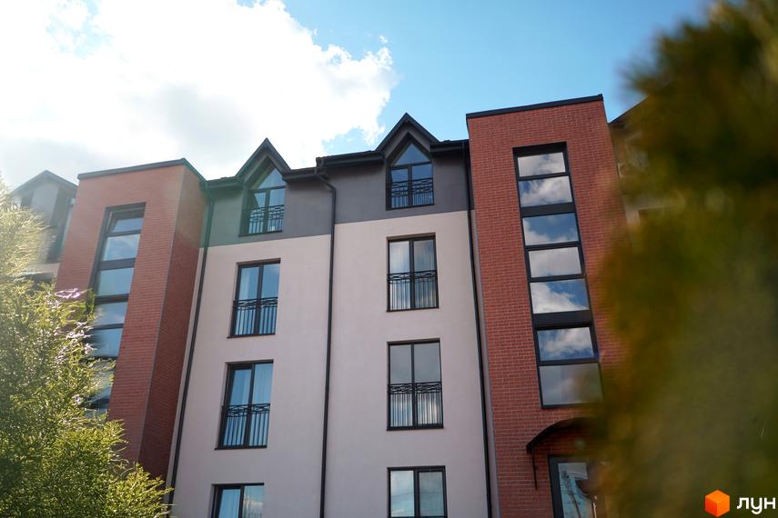 Фасад житлового комплексу Амстердам з цегляними та світлими елементами, великими вікнами та балконами. Навколо будівлі видно зелені дерева та блакитне небо з хмарами.