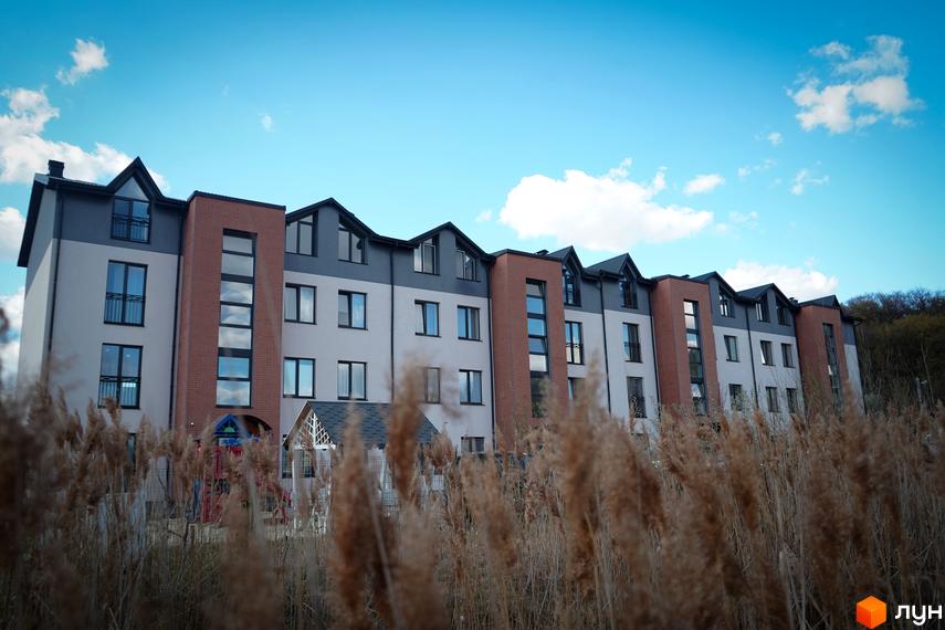 Зовнішній вигляд житлового комплексу Амстердам з цегляними та світлими фасадами, вікнами та балконами, на фоні блакитного неба з хмарами. На прибудинковій території видно дитячий майданчик.