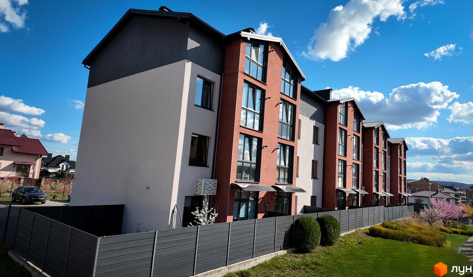 Житловий комплекс Амстердам – це цегляні будинки з великими вікнами та сірими фасадами, розташовані біля зеленої зони з кущами та деревами.