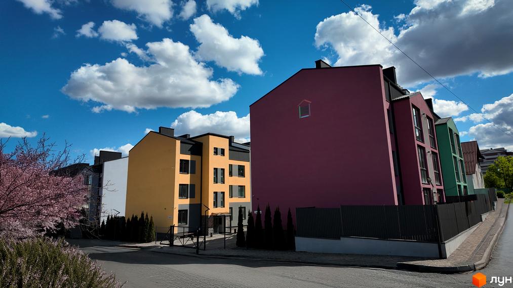 Житловий комплекс Вілла Італія – це сучасна новобудова з яскравими фасадами та доглянутою прибудинковою територією. На фото видно кольорові будинки під блакитним небом з хмарами.