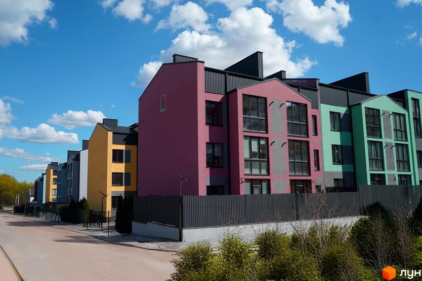 Комплекс ЖК Вілла Італія у яскравих кольорах фасаду з великими вікнами та сірим парканом. Навколо будівель зелені насадження та асфальтована дорога.