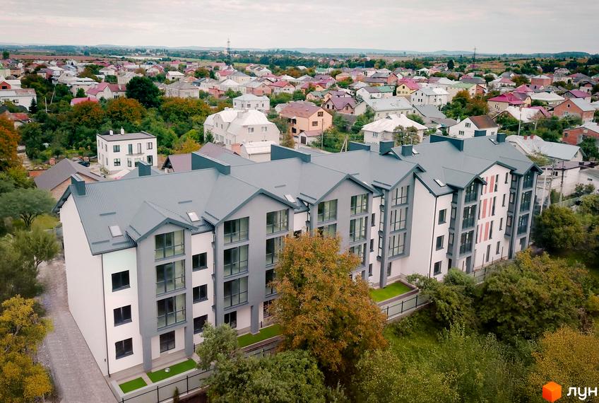 Зовнішній вигляд житлового комплексу Грушевського з сірими дахами та білими стінами, навколо зелені дерева та доглянута територія.