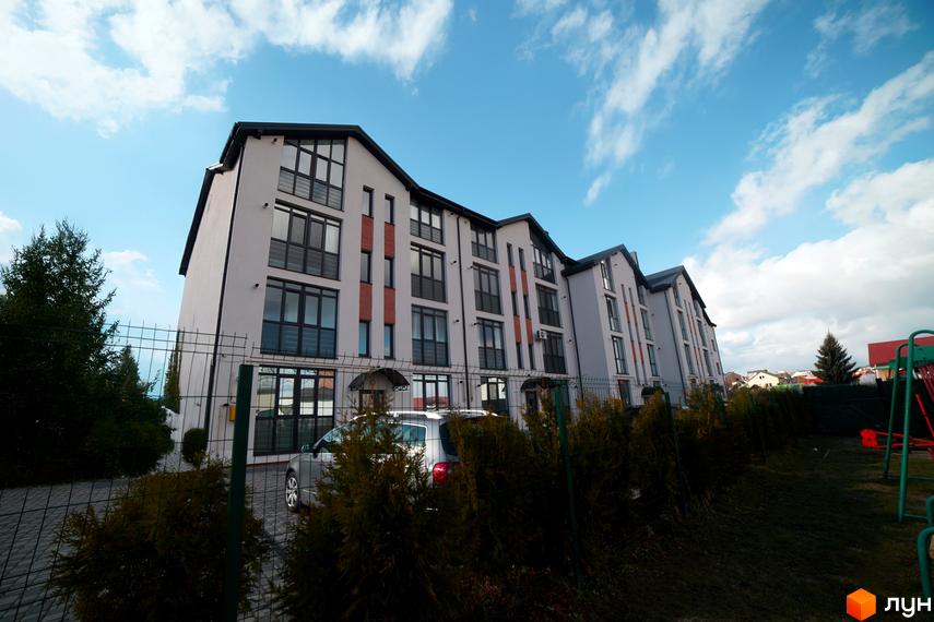 Зовнішній вигляд житлового комплексу Грушевського з численними вікнами та цегляними вставками. Прибудинкова територія включає парковку, зелені насадження та дитячий майданчик.