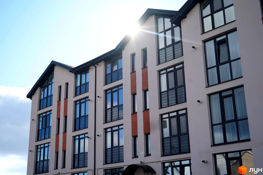 Фасад житлового комплексу Грушевського з великими вікнами та цегляними вставками під сонячними променями. Зовнішній вигляд новобудови з чіткими лініями та сучасною архітектурою.