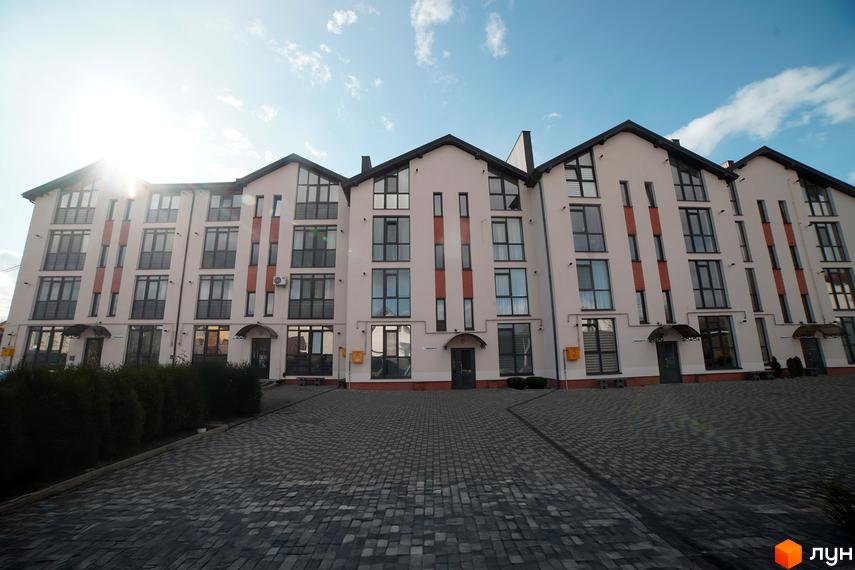 Зовнішній вигляд багатоквартирного будинку ЖК Грушевського з сірим дахом та світлими фасадами. Перед будинком розташована бруківка та зелені насадження.