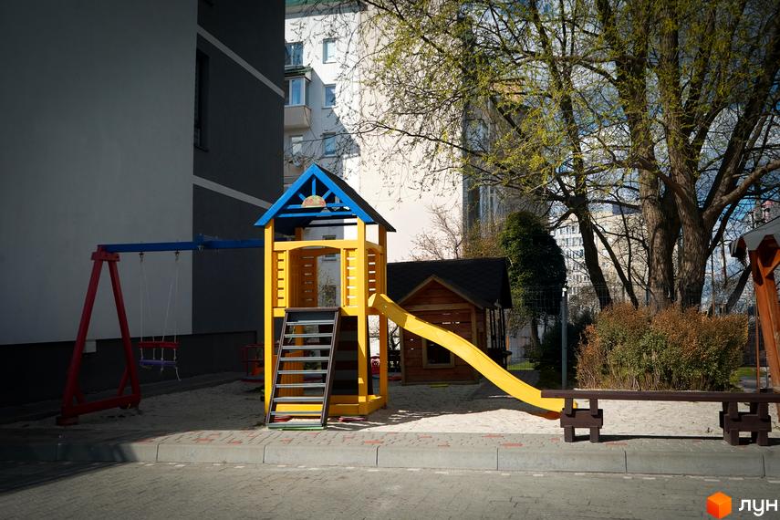 Дитячий майданчик біля житлового комплексу Флоренція з жовтим ігровим будиночком, гіркою та гойдалкою. Навколо пісок та зелені насадження.