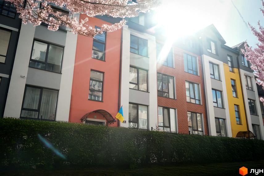 Житловий комплекс Флоренція з фасадами різних кольорів та вікнами, на фоні блакитного неба та гілок квітучих дерев. Біля будинку зелений газон.