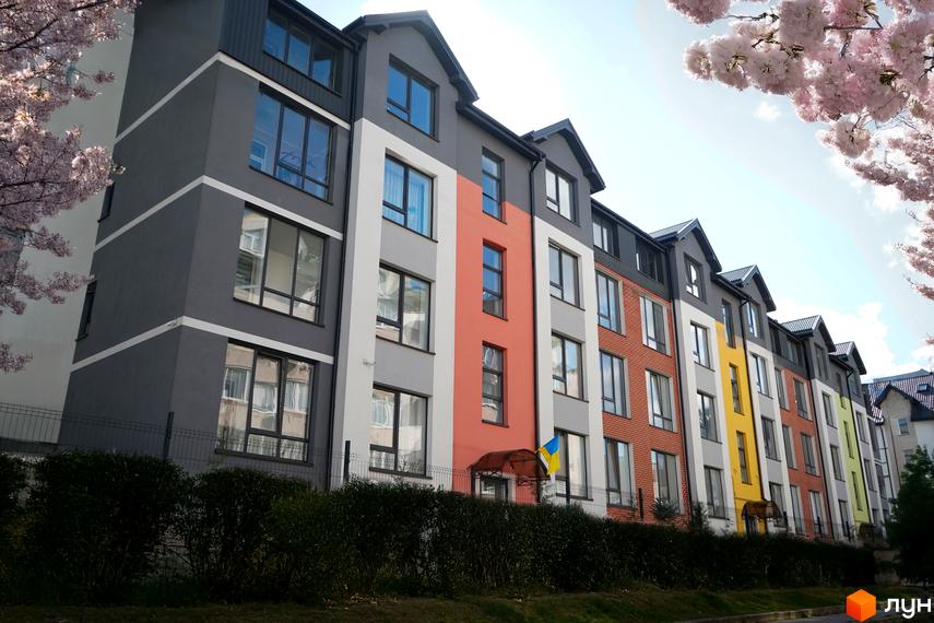 Житловий комплекс Флоренція - це багатоповерховий будинок з яскравим фасадом та вікнами, розташований на фоні блакитного неба та квітучих дерев. Перед будинком - зелені кущі та паркан.