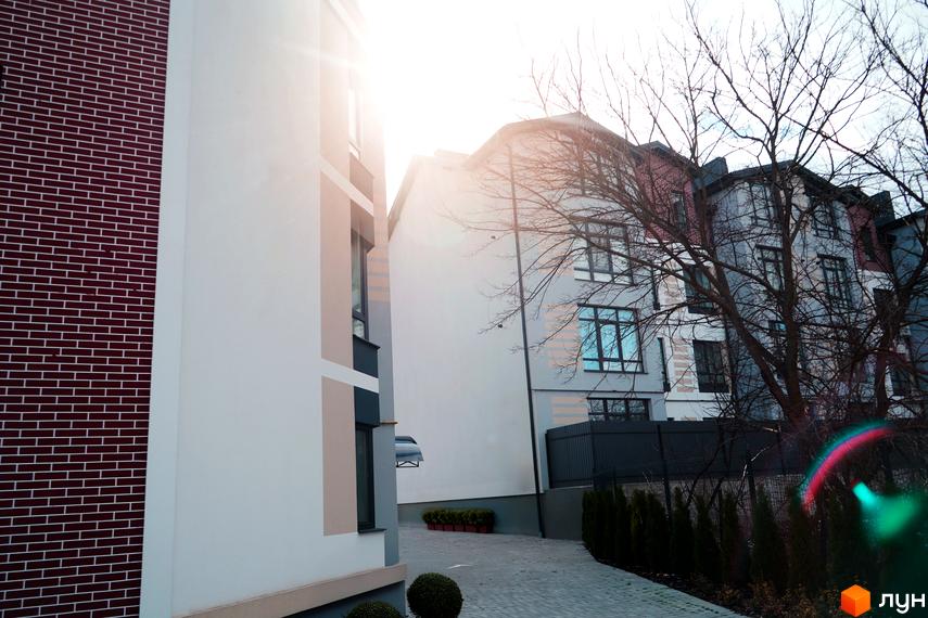 Зовнішній вигляд новобудови ЖК Вілла Венеція з цегляним фасадом та білими елементами, прибудинкова територія з деревами та доріжкою.