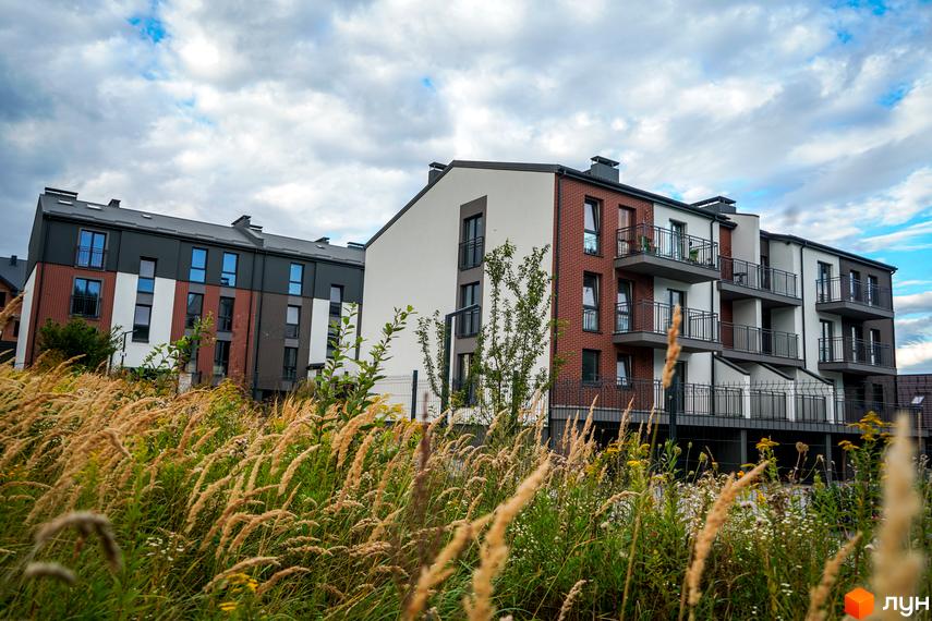 Житловий комплекс Вілла Швейцарія з цегляним фасадом та балконами на тлі хмарного неба. Попереду висока трава та зелені насадження.