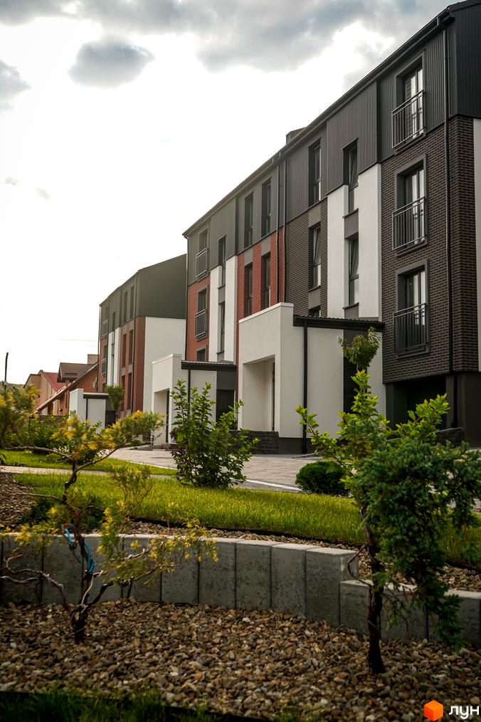 Зовнішній вигляд будинків ЖК Вілла Швейцарія з цегляним та білим фасадом, балконами та прибудинковою територією з газоном та деревами.