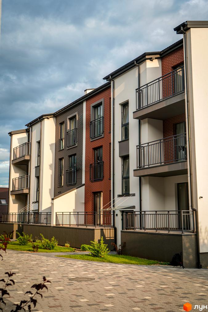 Зовнішній вигляд новобудови ЖК Вілла Швейцарія з цегляними та білими фасадами, балконами та доглянутою прибудинковою територією з травою та кущами.