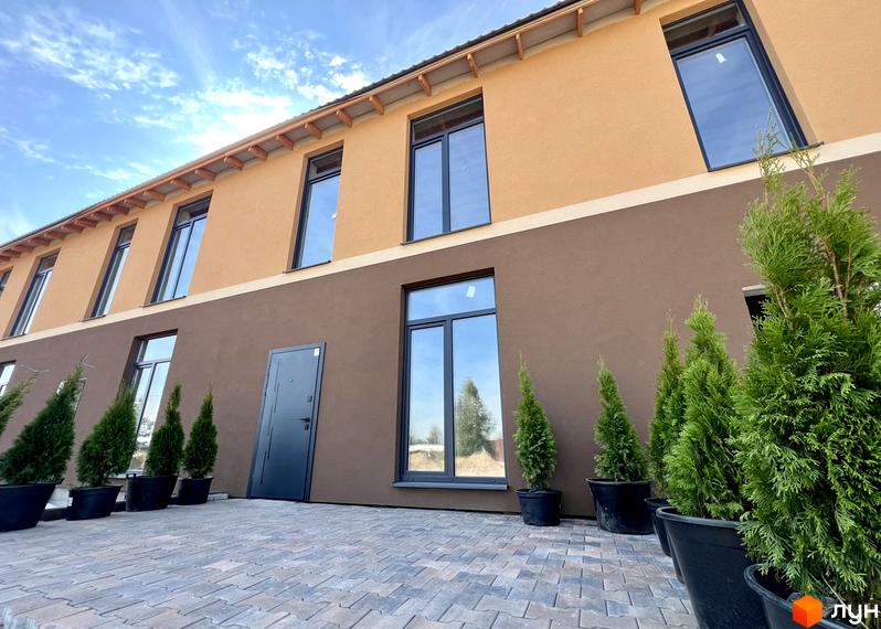 Фасад таунхаусів Лісовий затишок з коричневою та помаранчевою штукатуркою, великими вікнами та вхідними дверима. Поруч висаджені туї в горщиках на бруківці.