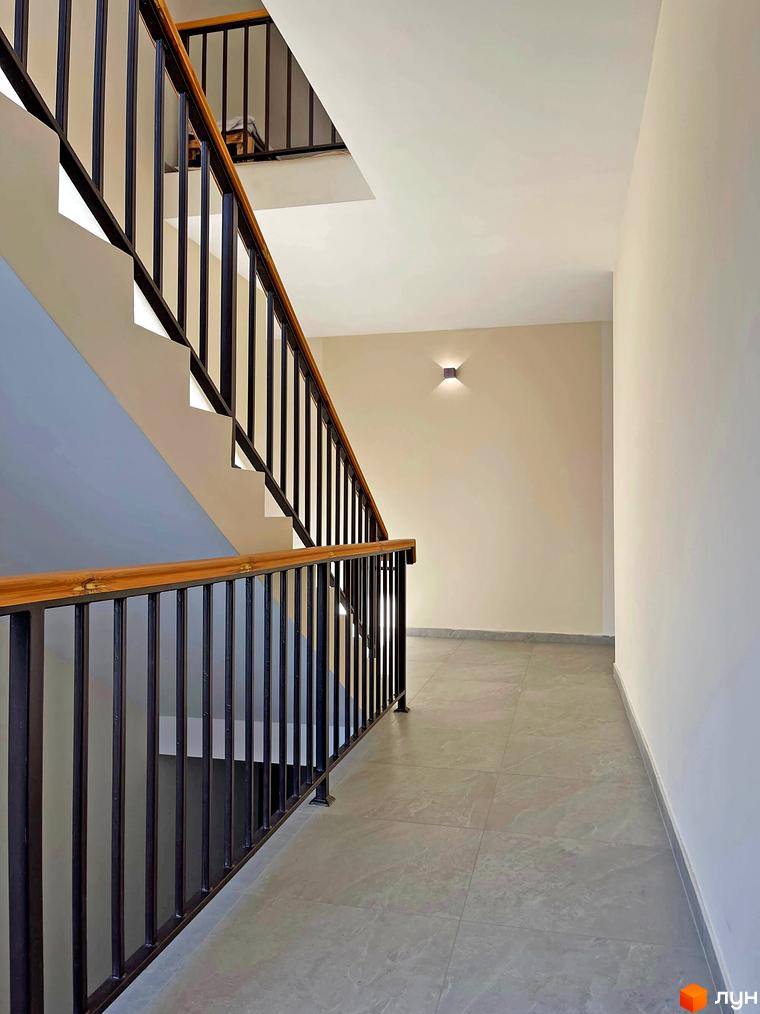 Сучасні сходи в житловому комплексі Smart Residence з дерев'яним поручнем та чорними металевими перилами. Підлога викладена плиткою.
