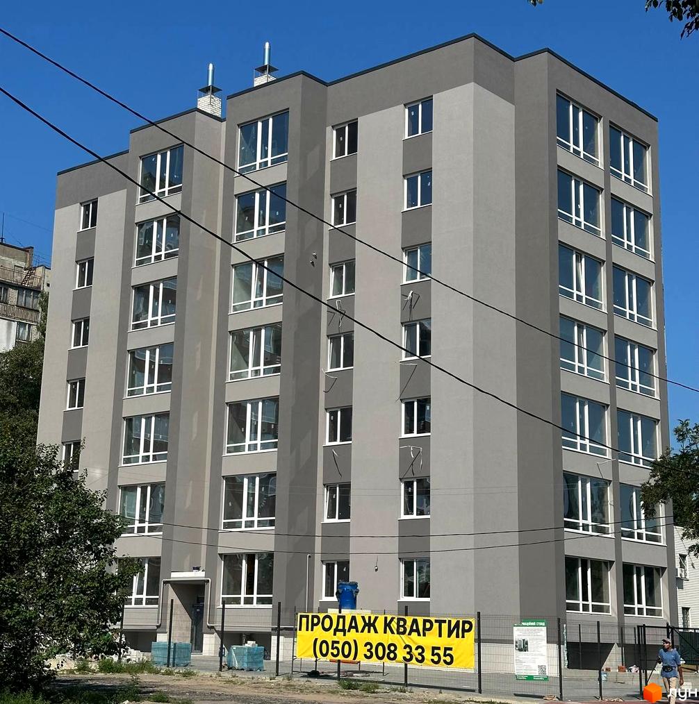 Сіра багатоповерхова новобудова з великими вікнами на тлі блакитного неба. Біля будинку огороджена територія з банером про продаж квартир.
