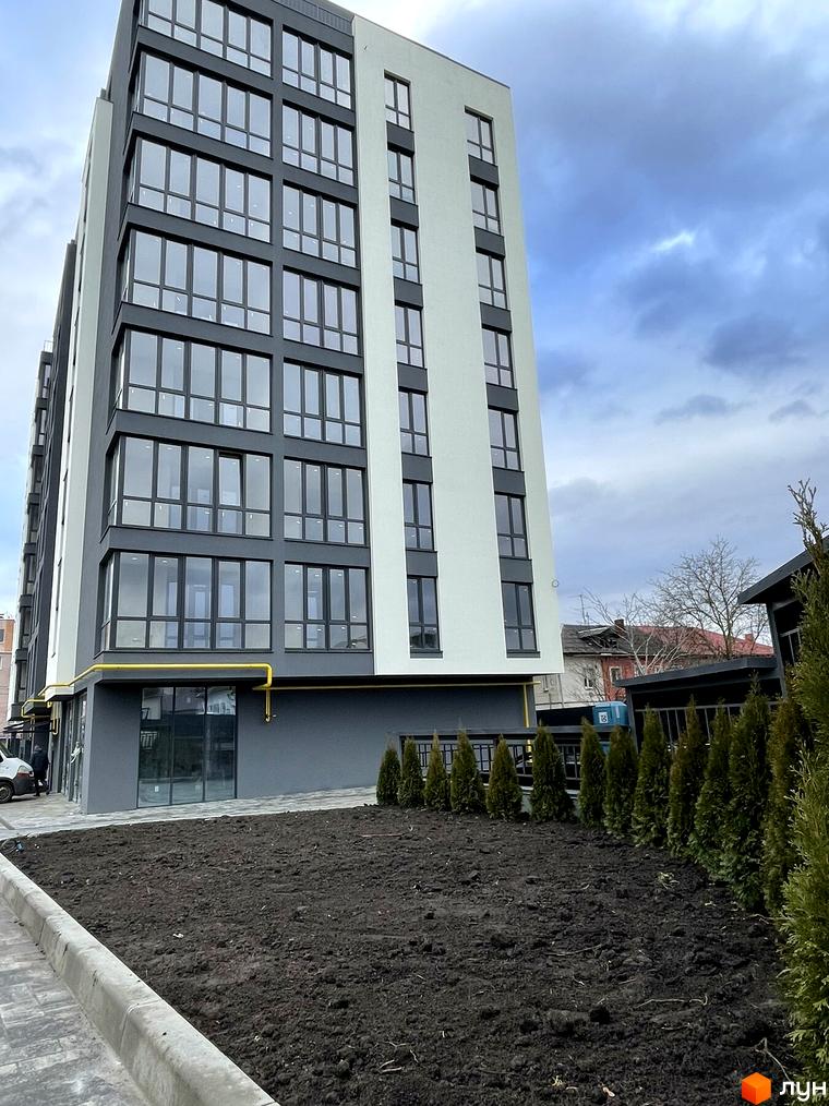 Зовнішній вигляд новобудови на вул. Будівельна, 1, з темними вікнами та світлою стіною, та прибудинковою територією з туями та чорноземом.