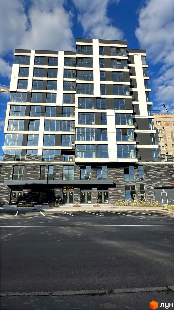 Сучасна новобудова з панорамними вікнами та темними фасадами, розташована біля парковки з розміткою.