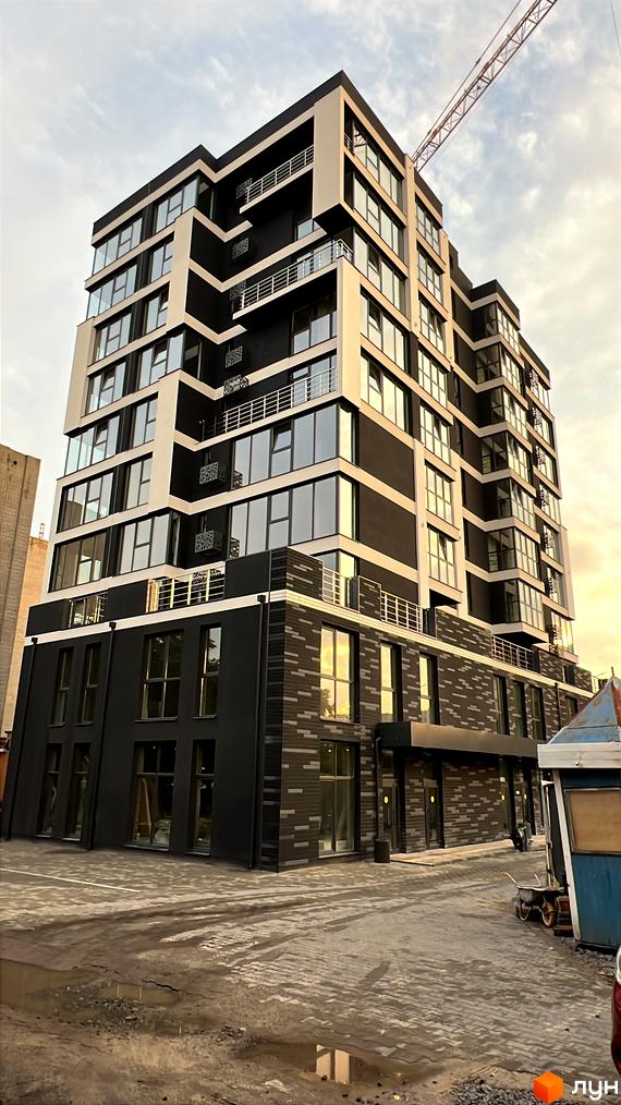 Зовнішній вигляд новобудови на вул. Родіона Скалецького, 29, з великою кількістю скління та балконами. Прибудинкова територія з бруківкою та будматеріалами.