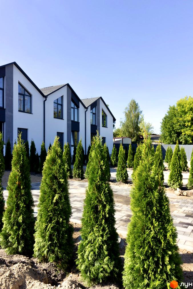 Таунхауси Small Town з біло-сірими фасадами та темними дахами, оточені зеленими туями та вимощеною територією. Зовнішній вигляд будинків та ландшафтний дизайн.