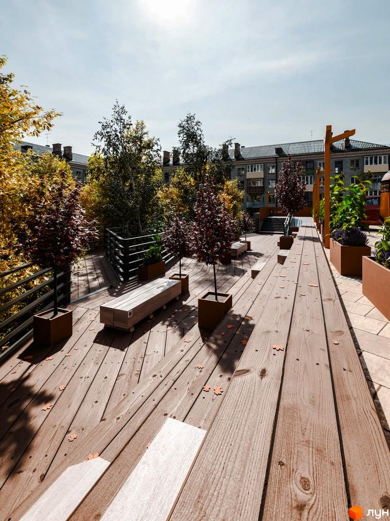 Житловий комплекс Перемога: тераса з дерев'яним настилом, лавками та деревами в горщиках. На задньому плані видно багатоповерхові будинки.