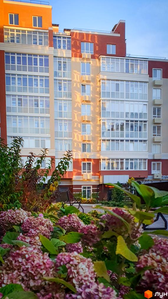 Новобудова Греків Ліс з квітучими рослинами на передньому плані. Фасад будинку оздоблений цегляними та світлими елементами, з великою кількістю скління.