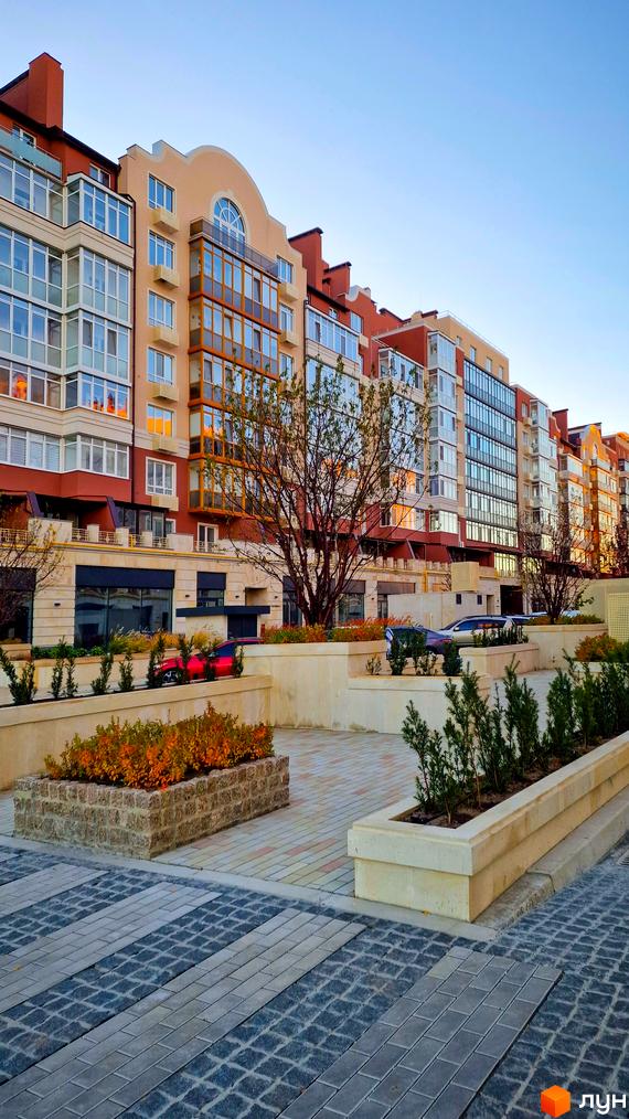 Житловий комплекс Греків Ліс з фасадами в бежевих та червоних тонах, скляними балконами та доглянутою прибудинковою територією з бруківкою та зеленими насадженнями.