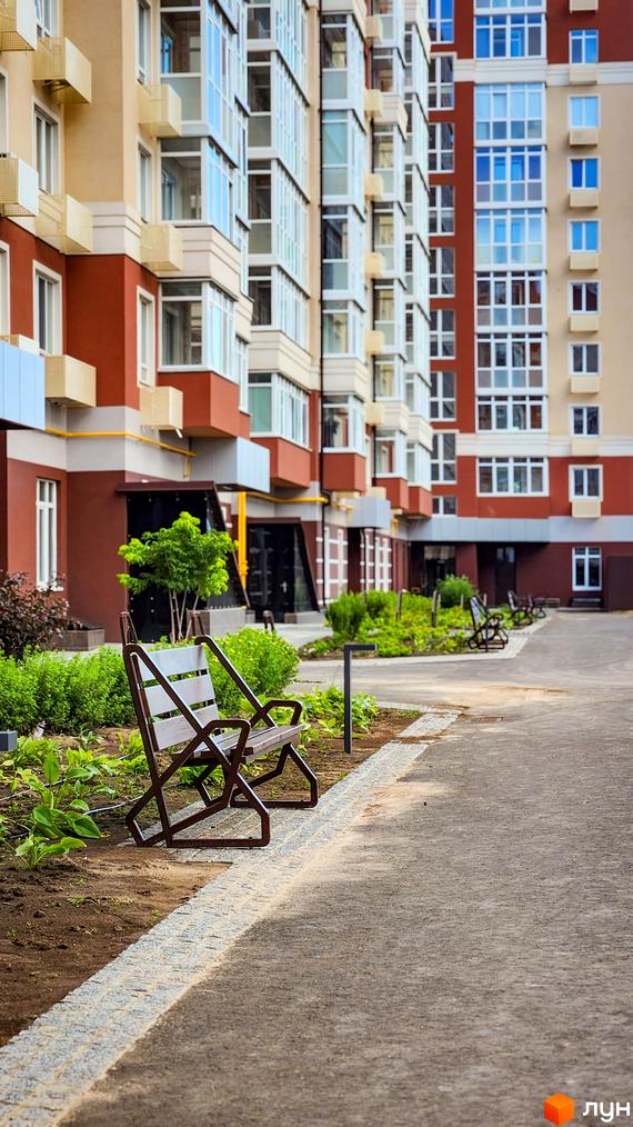 Фасад житлового комплексу Греків Ліс з балконами та вікнами, поруч розташована лавка та зелені насадження.