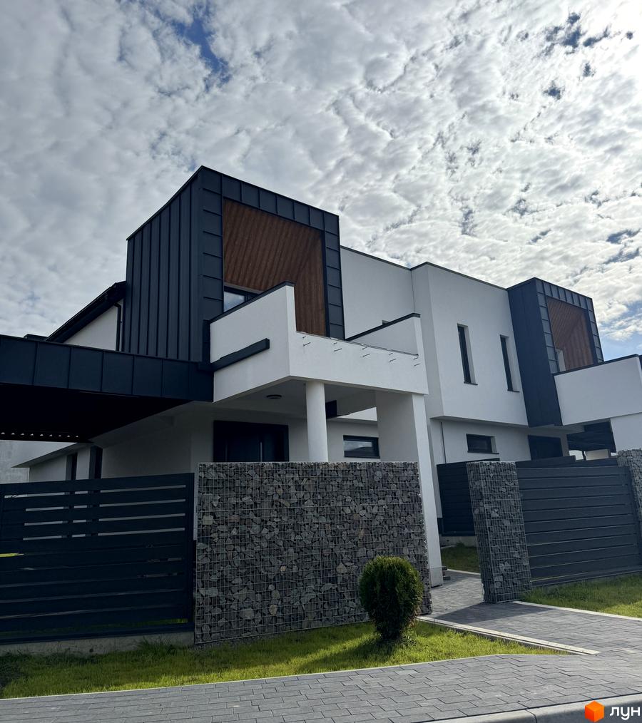 Котеджне містечко КМ Lake House Sokilnyky. Зовнішній вигляд будинку з білими стінами, темним дахом та дерев'яними вставками. Прибудинкова територія з парканом з каменю та металевими елементами.