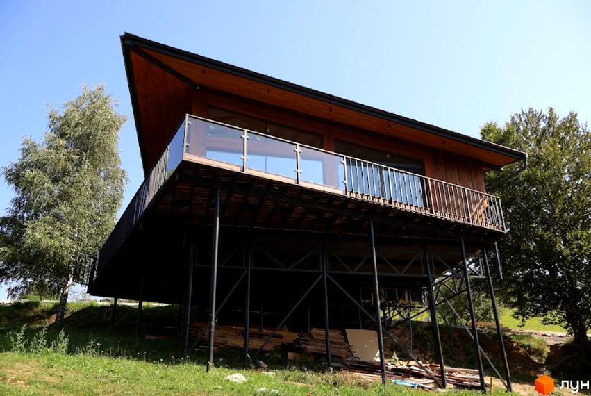 Котеджне містечко КМ Тишагора: дерев'яний будинок на палях з балконом та скляною огорожею, оточений деревами.