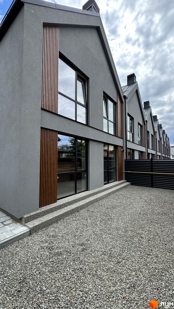 Зовнішній вигляд таунхаусів Legend Town з сірою штукатуркою та дерев'яними елементами. Перед будинками розташована гравійна прибудинкова територія та чорний паркан.