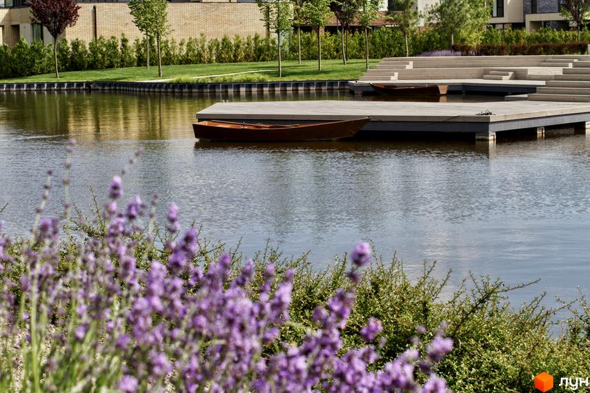 Котеджне містечко О.Краса з озером та дерев'яними настилами, оточене зеленню та квітами.