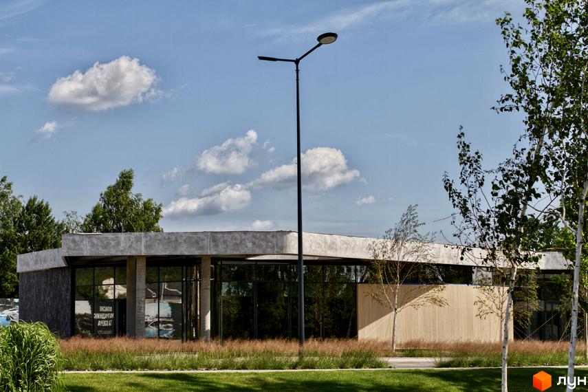 Котеджне містечко О.Краса: одноповерхова будівля з панорамними вікнами та бетонним оздобленням фасаду, оточена зеленню та деревами.