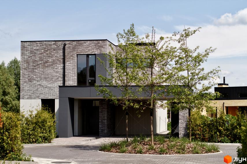 Котеджне містечко О.Краса: двоповерховий будинок з цегляним фасадом та великими вікнами, оточений зеленню та деревами. Прибудинкова територія з бруківки та ландшафтного дизайну.