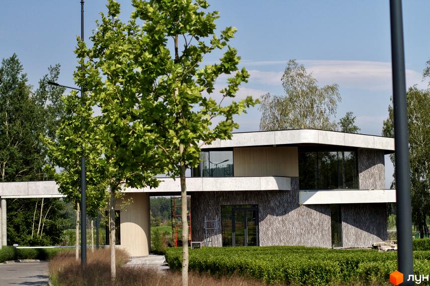 Котеджне містечко О.Краса: двоповерхова будівля з великими вікнами та сірим фасадом, оточена зеленню та деревами. Прибудинкова територія з доглянутими кущами та травою.