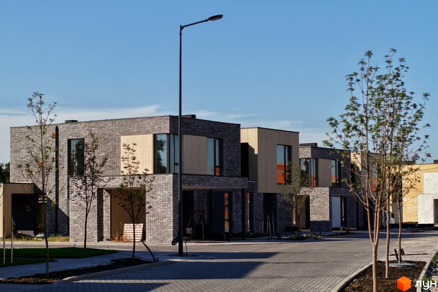 Котеджне містечко О.Краса: фасади будинків з сірої цегли та дерев'яних панелей, брукована дорога та зелені насадження.