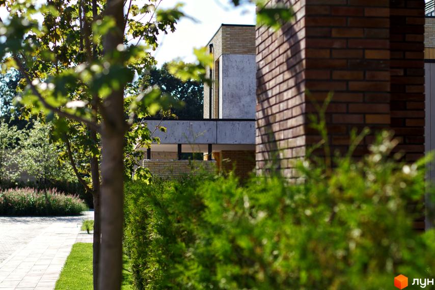 Котеджне містечко КМ О.Краса: цегляний фасад будинку та зелена прибудинкова територія з деревами та кущами.