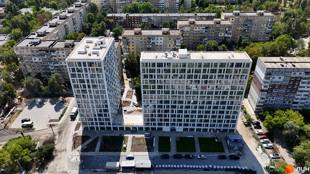 Житловий комплекс Вертикаль - це двосекційна новобудова з білими фасадами та численними вікнами, розташована поруч із багатоповерховими будинками та зеленими насадженнями.