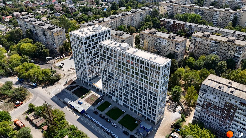 Новобудова ЖК Вертикаль з білим фасадом та численними вікнами, оточена деревами та дорогою.