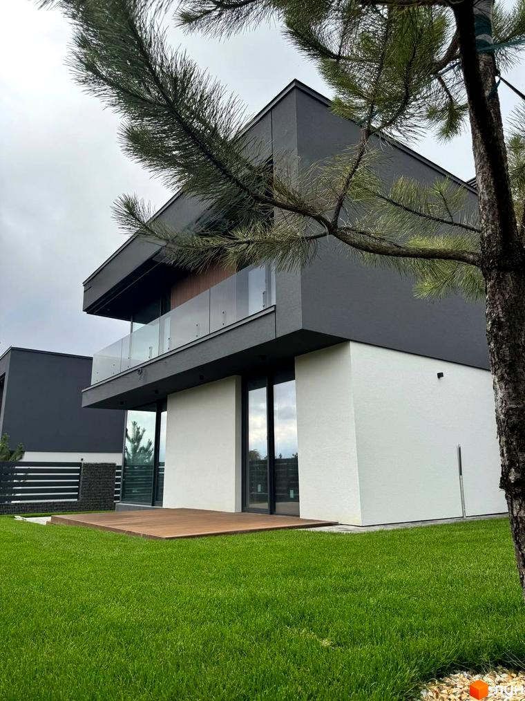 Зовнішній вигляд двоповерхового будинку в Котеджному містечку Prime Hill з великими вікнами та дерев'яною терасою на зеленій галявині.