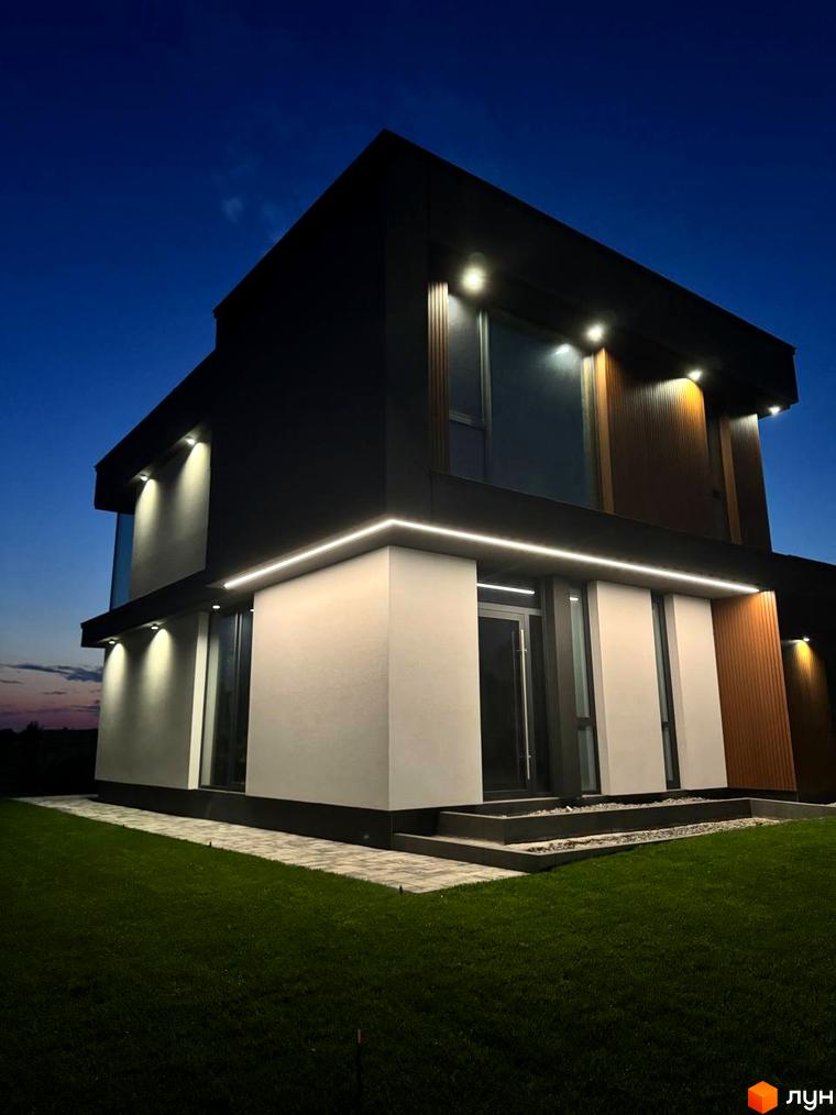 Зображення двоповерхового будинку в Котеджному містечку Prime Hill з підсвічуванням фасаду та доглянутим газоном на прибудинковій території.