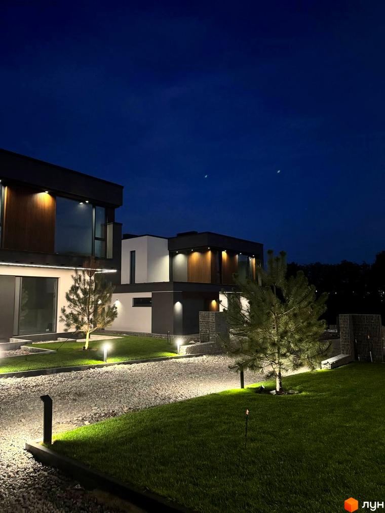 Зображення Котеджного містечка Prime Hill вночі: освітлені будинки з великими вікнами та доглянута територія з газоном і декоративними деревами.