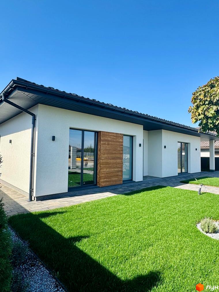 Зображення будинку в Котеджному містечку Bratske Home з великими вікнами та дерев'яною вставкою. Навколо будинку зелений газон та доріжки з плитки.