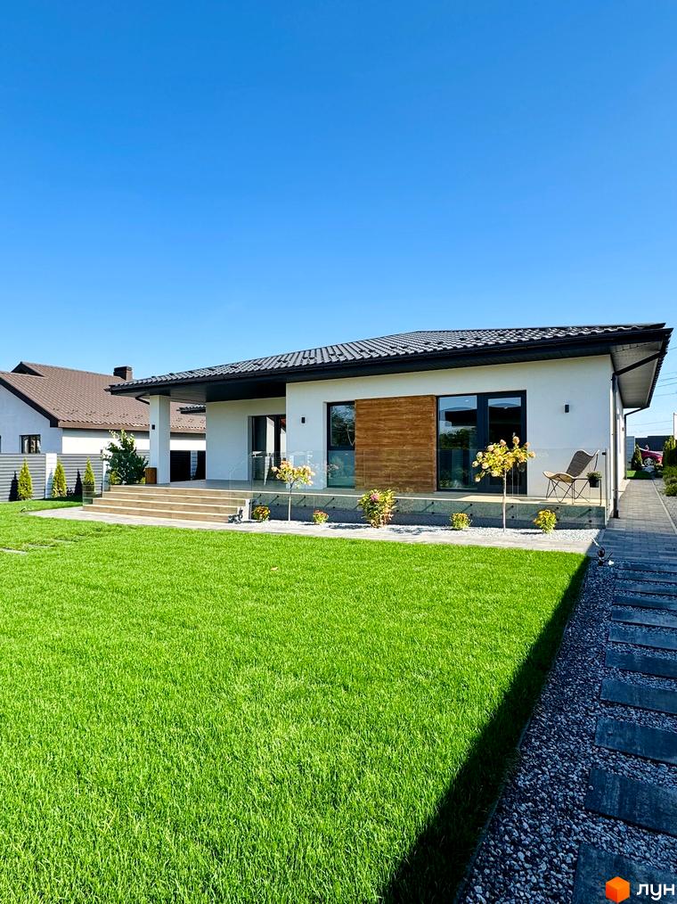 Вигляд будинку в Котеджному містечку Bratske Home з великим газоном та доріжкою. На території висаджені декоративні дерева та квіти.