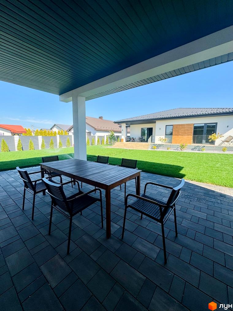 Вид на будинок з терасою та меблями в Котеджному містечку Bratske Home. Навколо будинку зелений газон і дерева.