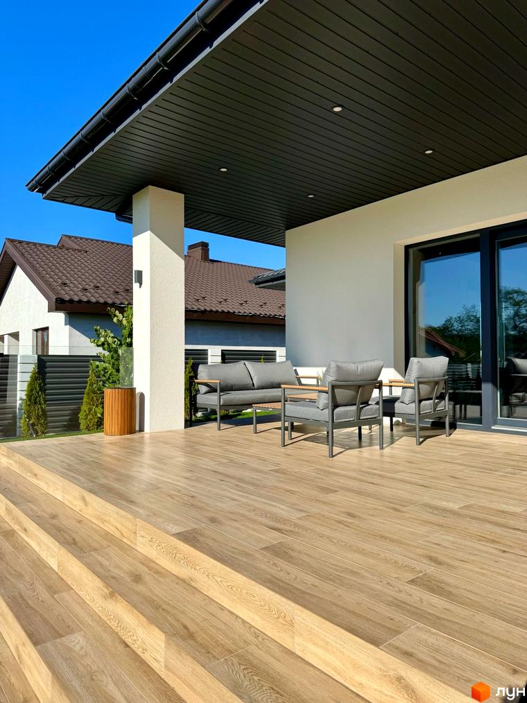 Зображення тераси Котеджного містечка Bratske Home з меблями для відпочинку. Вигляд на будинок з коричневим дахом та зелений газон.