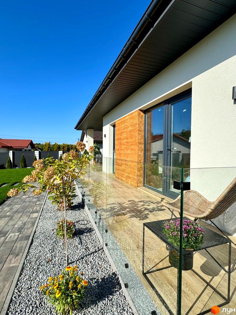 Зображення будинку з терасою в Котеджному містечку Bratske Home. Навколо будинку розташовані декоративні рослини та каміння.