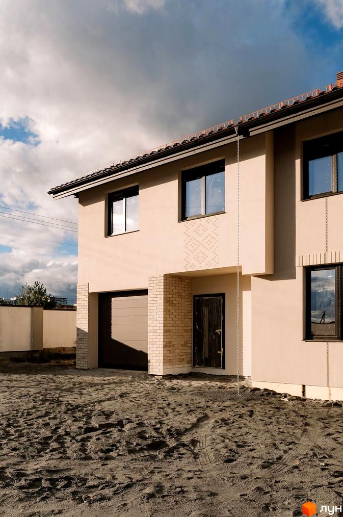 Зображення будинку в Котеджному містечку Етнія. Двоповерховий будинок з гаражем та цегляним оздобленням, перед будинком піщаний ґрунт.