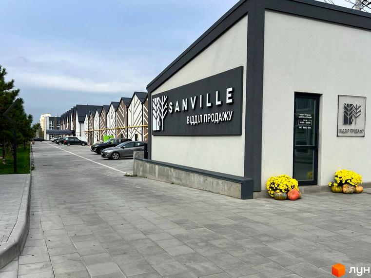 Фасад будинку відділу продажу Житлового комплексу Sanville Park з вивіскою та осіннім декором. Навколо розташована доглянута територія з тротуарною плиткою.