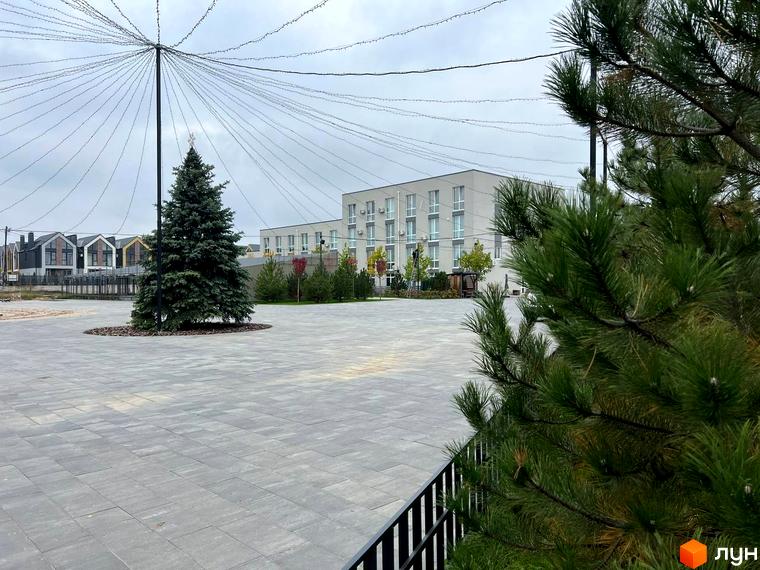 Зображення площі з ялинкою та будівлею в Житловому районі Sanville Park. На передньому плані зелені сосни та чорна огорожа.