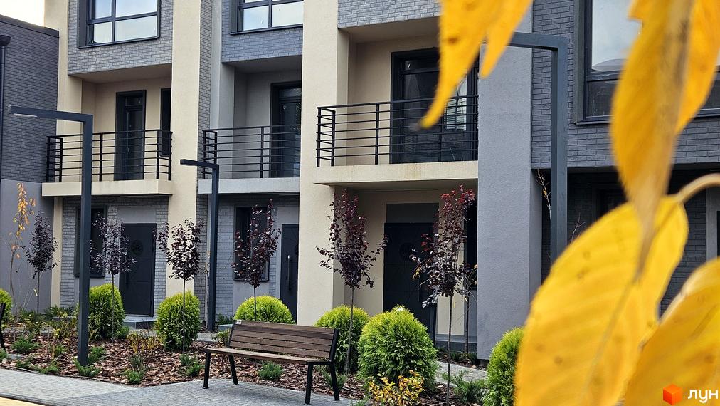 Фасад таунхаусів Sвій Dім 2 з балконами та чорними дверима. На території ландшафтний дизайн з деревами та лавкою.